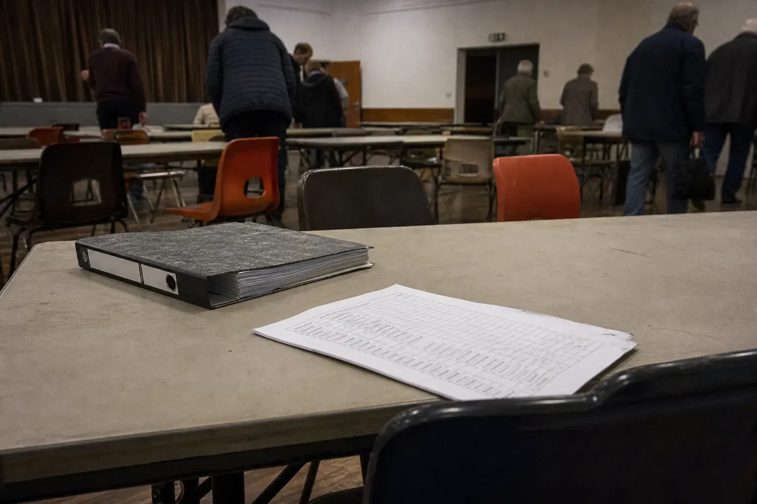 Salle polyvalente municipale en fin de réunion, tables pliantes, chaises dépareillées, un classeur et une feuille de…