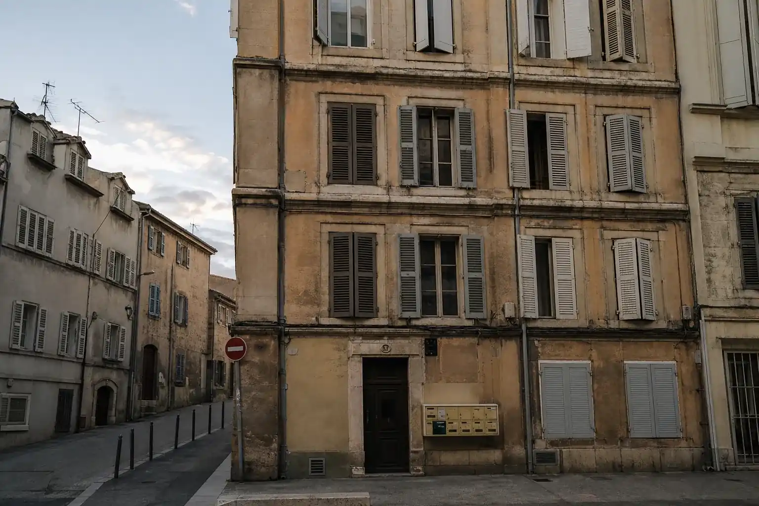 Façade d’immeuble ancien de rapport, cadrage frontal légèrement décentré, fin de journée, volets hétérogènes, boîtes…
